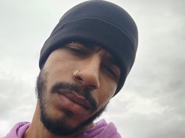 AfroJake con un berretto nero, una felpa viola, un anello al naso e una barba, che guarda leggermente di traverso con un'espressione compiaciuta. Cielo nuvoloso sullo sfondo..