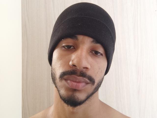 AfroJake con un berretto nero, occhi marroni, un piccolo anello al naso e una barba nera, che guarda leggermente di lato con un sorriso appena accennato. Senza camicia, sfondo in legno..