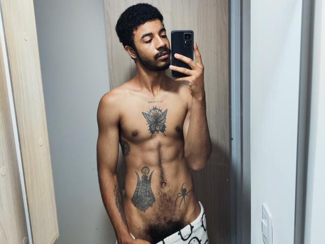 AfroJake, sin camisa, con cabello negro rizado, sosteniendo un teléfono, mostrando tatuajes en el pecho y los abdominales, usando pantalones blancos, de pie en un selfie frente a un espejo, mirando a la cámara..
