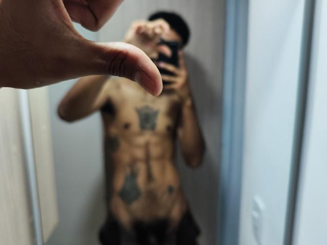 De getatoeëerde, gespierde torso van AfroJake, met een opvallende tatoeage op de borst, is wazig in de spiegel te zien. Een hand wijst in de voorgrond naar de camera. De zwarte broek van AfroJake is zichtbaar..
