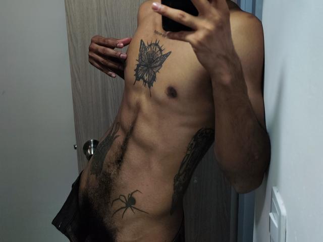 Le torse musclé et bronzé d'AfroJake, avec un tatouage de papillon sur la poitrine, un tatouage d'araignée sur le bas de l'abdomen, et une main couvrant son visage dans une photo de lui-même devant un miroir..