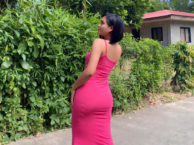 BettinaJohnson stands in a pink dress, showing her big ass, with short black hair, looking back, in a green leafy backyard, on a wet sidewalk.