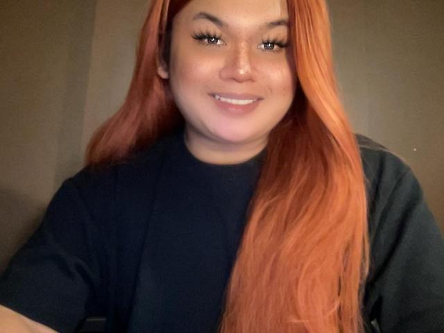ThaniaMayLovez with long red hair, black shirt, smiling, freckled face, and eyeliner, looking at the camera. Her tits are not visible. Her ass is not visible.