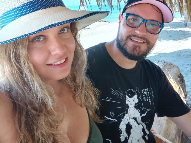 MsMiaMrSergio with wavy blonde hair, big boobs in a green top, and a sunhat, next to bearded MrSergio in a black Batman shirt and pink cap.