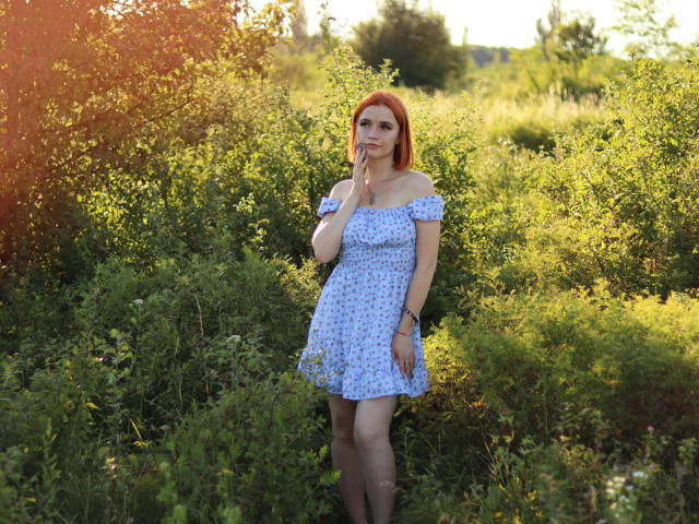 MargaritaKis with red hair, wearing a blue floral off-shoulder dress, stands in green bushy area, touching her chin, sunlight filtering through leaves, her ass slightly visible.
