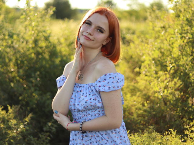 MargaritaKis with red hair, freckles, and a cute smile, wearing an off-shoulder blue floral dress, touching her neck, standing in a sunlit green field.