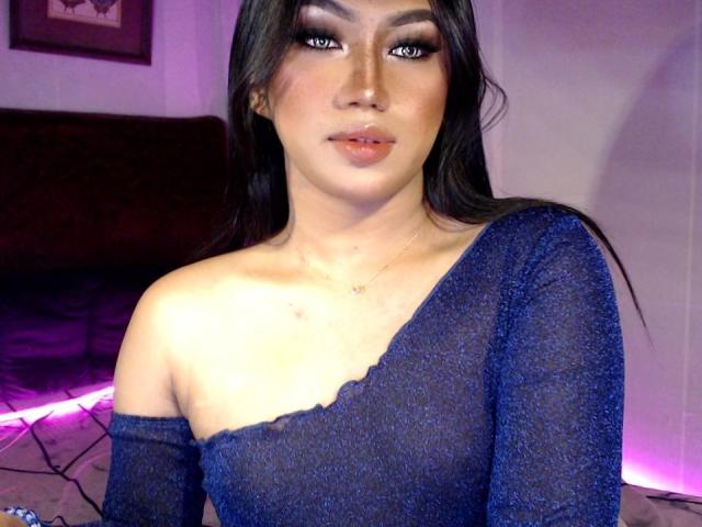 GoddessXsteffie with long black hair, blue glitter off-shoulder dress, and noticeable makeup, has medium-sized tits and a cute face, looking at the camera with purple-lit background.
