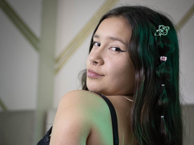 LilithSwett has dark green hair with a pink butterfly hairpin, black eyeliner, a nose ring, and a lip piercing. She's looking back with a slight smile. Boobs partially visible.