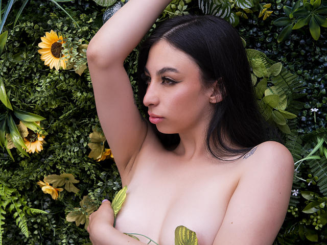 CamilleRios, nude with small tits, dark hair, arm raised, green leaf covering her pussy, surrounded by sunflowers and foliage, looking down with a serious expression, soft light.