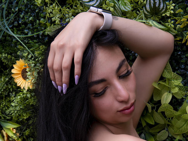 Camille Rios, met lang zwart haar, paarse nagels en een roze horloge, met gesloten ogen, hand op haar hoofd, een zonnebloem aan de linkerkant, een groene, bladerrijke achtergrond. Haar achterkant is niet zichtbaar..