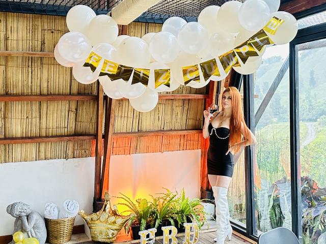 Leared with long red hair, black dress, and white thigh-high boots, stands holding a microphone, surrounded by white balloons and gold letters, singing at a party.
