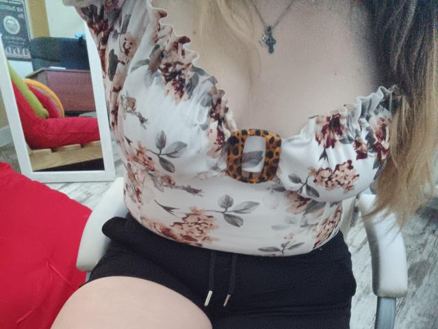 Leiliia has small tits, wearing a floral top with a leopard print button, black shorts, and a necklace, sitting on a white chair, reflected in a mirror.