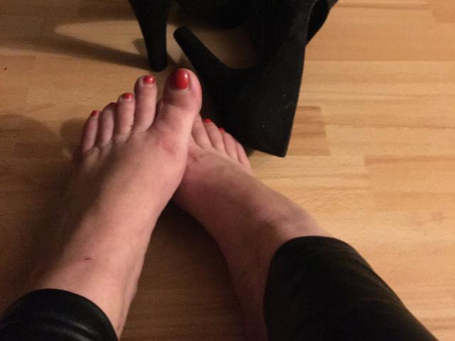 Lady-Kim-hot's feet with red-painted toes, slightly bruised, crossed on a wooden floor, black boots in the background.