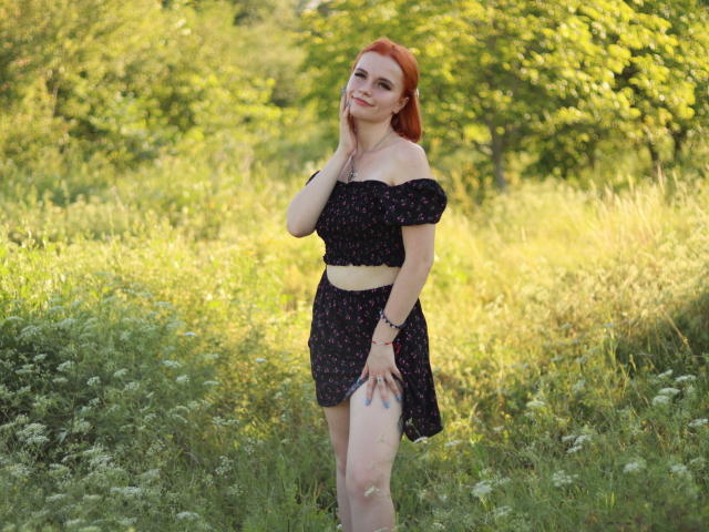 MargaritaKis with red hair, black off-shoulder top, and matching skirt, standing in a grassy field, touching her cheek, smiling, showing her pale, toned ass.