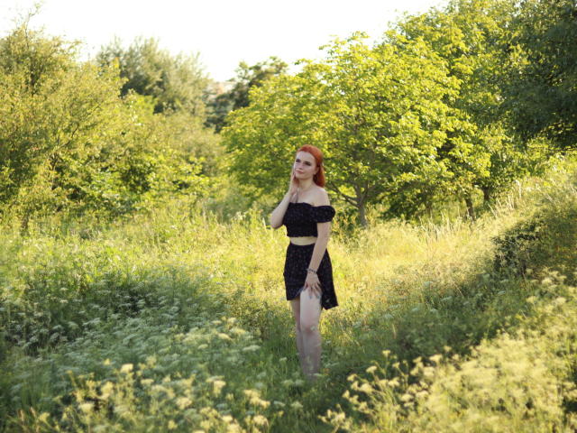 Margarita Kis steht auf einer sonnendurchfluteten, grasbewachsenen Fläche, trägt ein schwarzes Kleid mit einem nackten Schulterteil, ihr rotes Haar bildet einen Kontrast zum grünen Laub, und sie hält eine Hand vor ihr Gesicht..