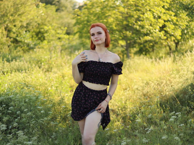 MargaritaKis hat rote Haare und trägt ein schwarzes, schulterfreies Oberteil mit Blumenmuster und einen passenden Rock. Sie steht auf einer grünen Wiese und hebt ihren Rock leicht an, um ihren nackten Oberschenkel zu zeigen..