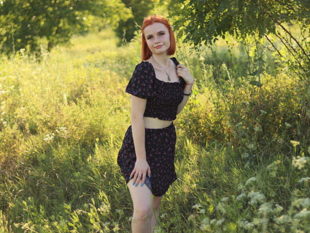 MargaritaKis with red hair, wearing a black floral crop top and skirt, standing in a grassy field, touching her hair, smiling, showing her ass, in sunlight.