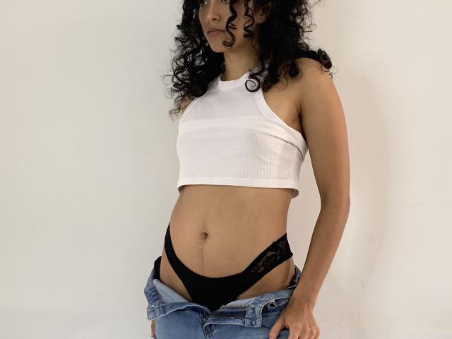 Vivienne Laurent, con cabello negro rizado, vestía un top corto blanco con textura, ropa interior de encaje negra y jeans azules desabrochados, mostrando su abdomen plano y sus glúteos pequeños..