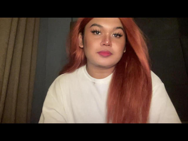 ThaniaMayLovez with long, red hair, white shirt, pink lipstick, and full eyebrows, looking directly at the camera with a neutral expression.