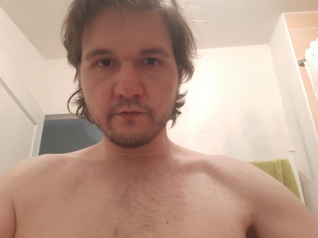 NectarineFad, shirtless, with messy brown hair, a beard, and chest hair, looking into the camera. His eyes are slightly down, and his expression is neutral.