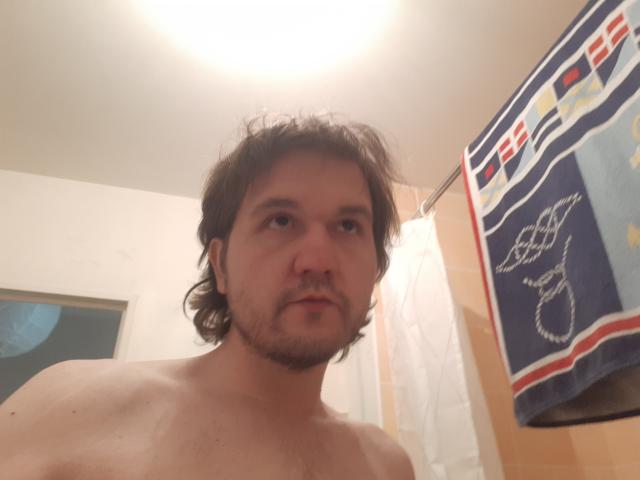 NectarineFad is a shirtless dude with messy brown hair and a beard, standing in a bathroom with a towel hanging on the right. His face looks bored.