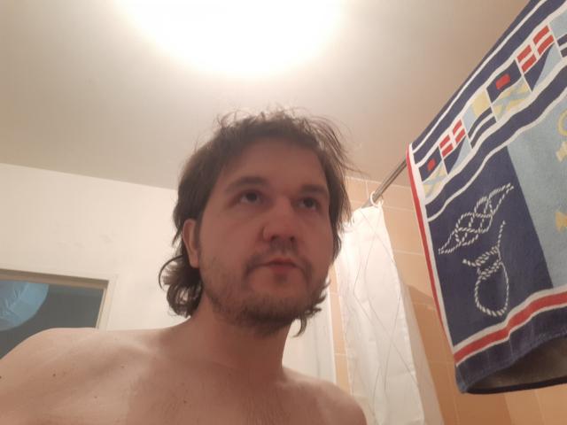 NectarineFad, shirtless, with messy brown hair and a beard, standing in a bathroom, looking up at the camera. Towel with a snake design on the right.