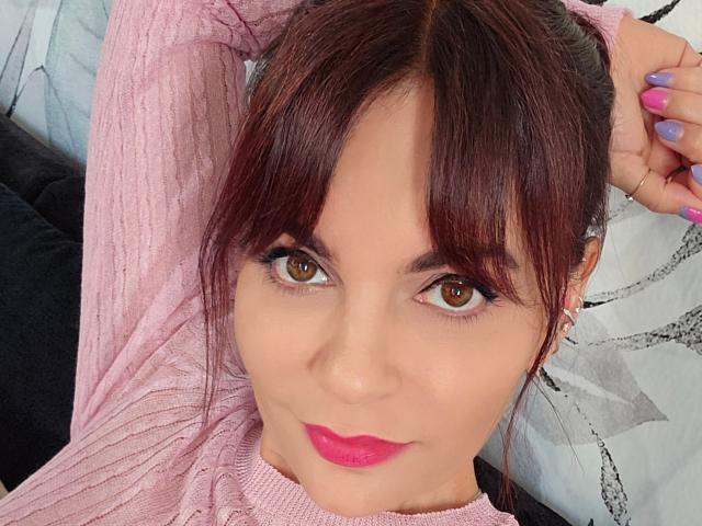 JuneLaFrenchie with brown eyes, pink lipstick, and dark brown hair in a ponytail, wearing a pink ribbed sweater, has purple nails, and small hoop earrings.