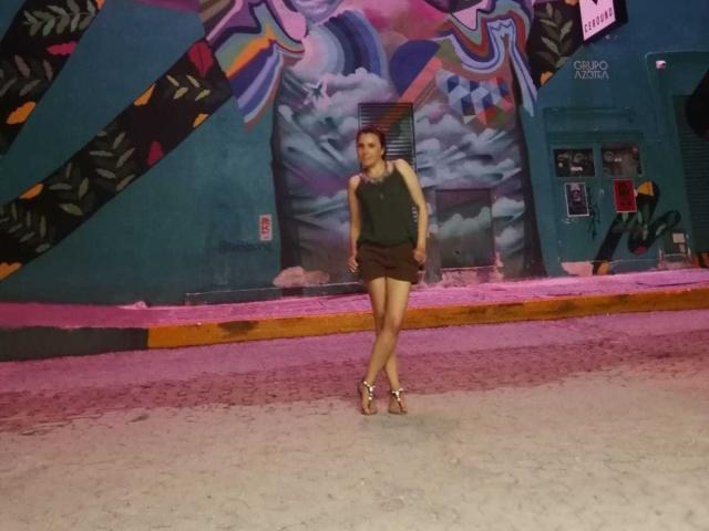 AdelaShery stands in a gritty urban setting, wearing a green tank top and brown shorts, with sandals, her ass slightly raised, in front of colorful graffiti.