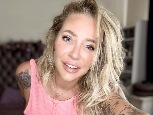 GlamSadie has blonde, wavy hair, blue eyes, pink lipstick, and tattoos on her arm. She's wearing a pink, ribbed tank top, smiling with her teeth visible.