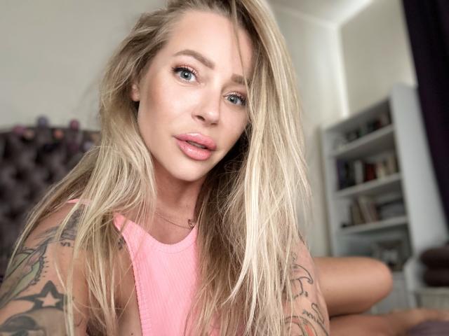 GlamSadie with blonde hair, blue eyes, pink lips, and tattoos, wearing a pink tank top, looking at the camera with a slight smile. Boobs visible, arms tattooed.