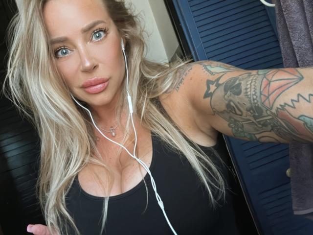 GlamSadie with blue eyes, long blonde hair, wearing a black top, white earbuds, and tattoos on her right arm, showing her boobs slightly. She's looking at the camera.