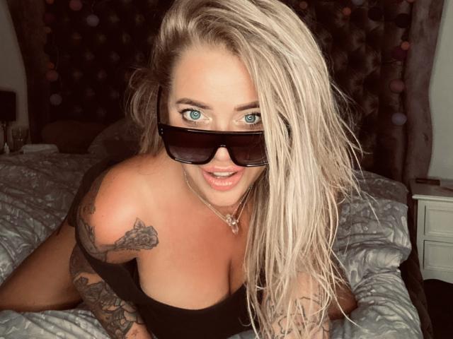 GlamSadie with blonde hair, blue eyes, black sunglasses, and tattoos, wearing a black top, showing her big boobs, and a necklace, leaning on a bed.