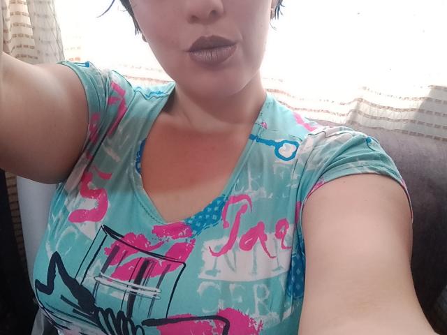 Valen69x with short black hair, wearing a teal shirt with pink and blue kiss prints, showing her tits and pouty, dark purple lips, holding a camera.