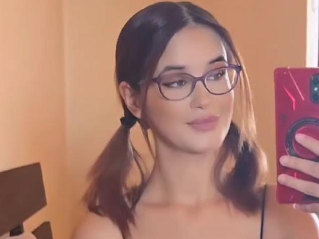 KoryHolly, com maria-chiquinhas, óculos e batom rosa, segurando um telefone vermelho, sorrindo, mostrando seus ombros nus e um rosto adorável. Seios não visíveis..
