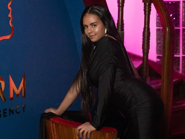 ArielleWilde with long black hair, wearing a tight black dress, leaning on a wooden staircase, showing her ass, looking back with a slight smile, pink light in background.