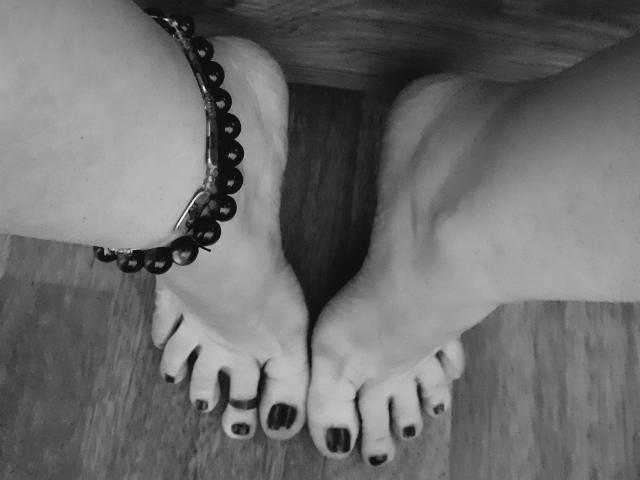 MindyTaylor's bare feet with dark toenail polish, wearing a bead bracelet on her right ankle, standing on a wooden floor.