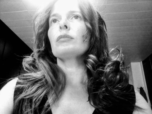 Black-and-white photo of MindyTaylor with wavy, shoulder-length hair, gazing upward, wearing a sleeveless top, showing her tits slightly, in a dimly lit room.