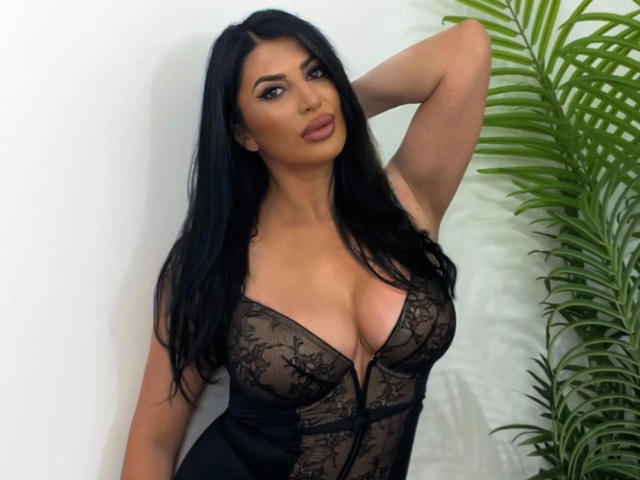 MayaCaprice trägt schwarze Spitzendessous, zeigt große Brüste und ein verführerisches Gesicht, hat langes, schwarzes Haar, einen Arm angehoben und steht neben einer grünen Pflanze..
