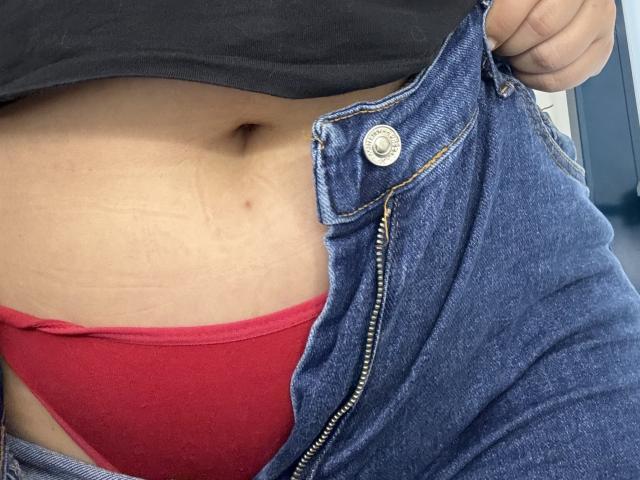 Barbarica's belly button, showing a slight tan line and a small mole, with blue jeans unzipped to reveal red panties, black shirt lifted.