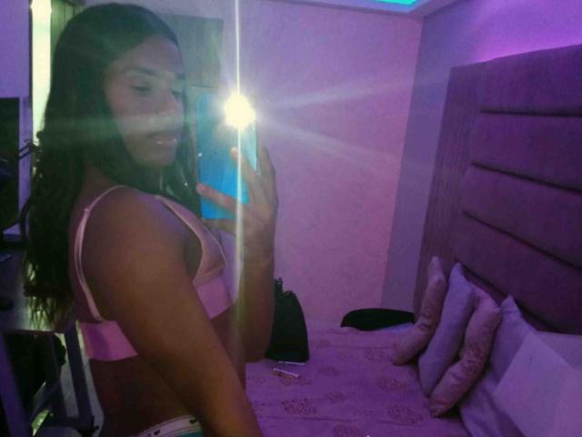 DanielisValen, con lunghi capelli scuri, reggiseno rosa e biancheria intima bianca, tiene in mano un telefono, riflesso nello specchio, flash, stanza viola, letto con cuscini, luce blu, sorridente, formosa, attraente..
