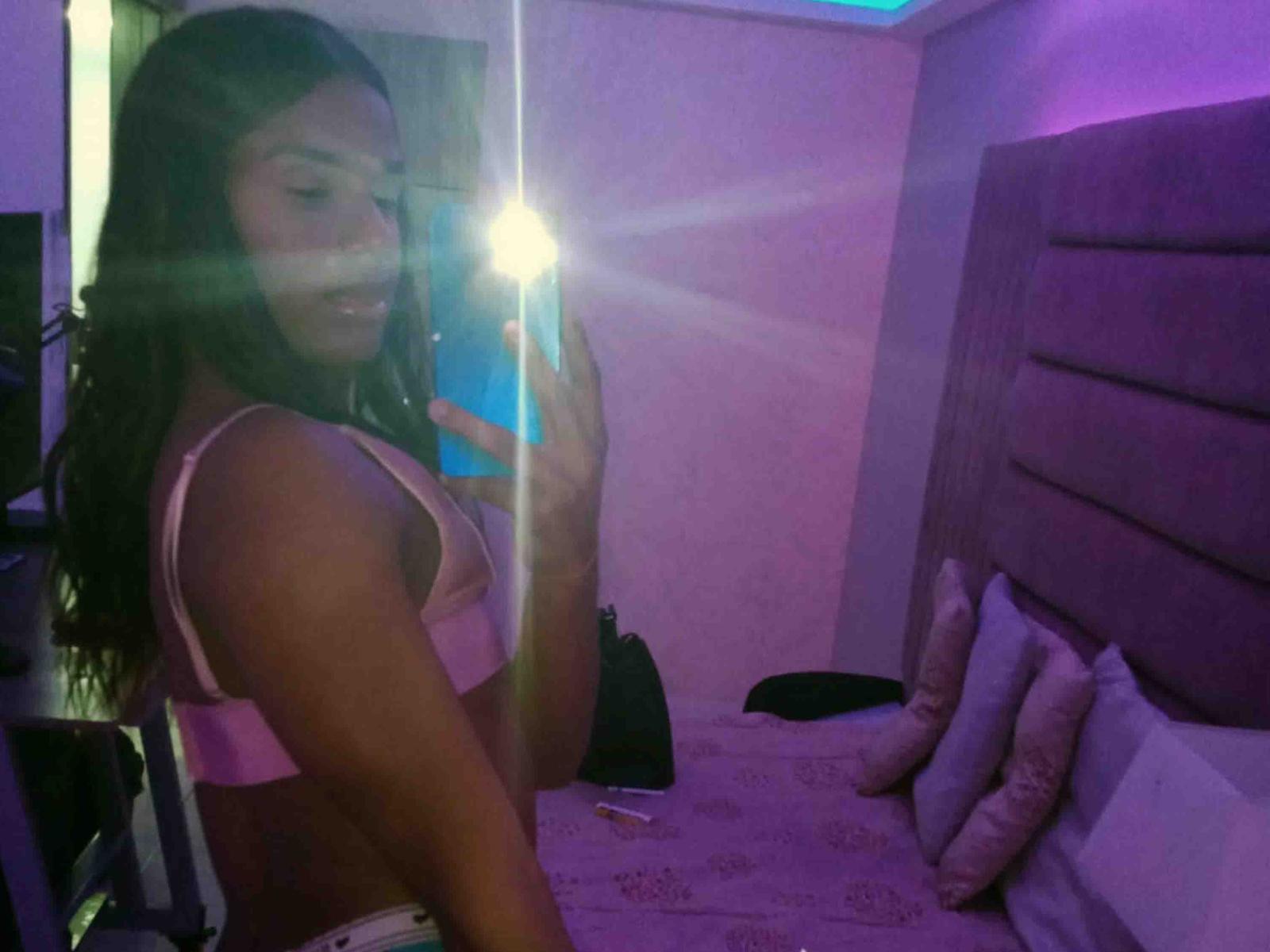 DanielisValen with long dark hair, pink bra, and white panties, holding a phone, mirror reflection, flash, purple room, bed with pillows, blue light, smiling, curvy, attractive.