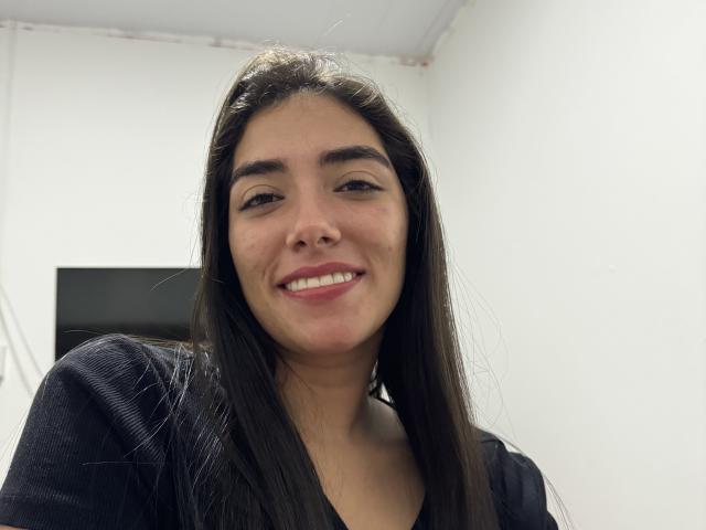 SharonKlart with long dark brown hair, smiling with pink lipstick, wearing a black top, her nose slightly red, in a bright room.