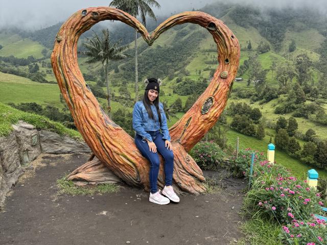 SharonKlart sits on a large, carved wooden heart, wearing a blue jacket, black beanie, and white sneakers, with her ass resting on the heart's base.