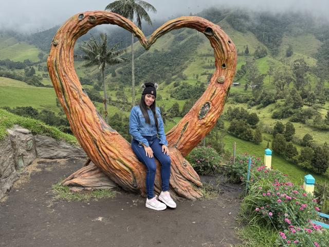 SharonKlart, met lang zwart haar, een blauwe jas, jeans en witte sneakers, zittend op een houten sculptuur in de vorm van een hart, met een zwarte hoed op, in een mistig, groen berglandschap..