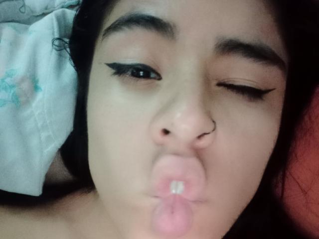 StellaBeckker with black eyeliner, winking, and pouting her pink lips with a nose ring, making a kissing face. She has dark hair and a light skin tone.