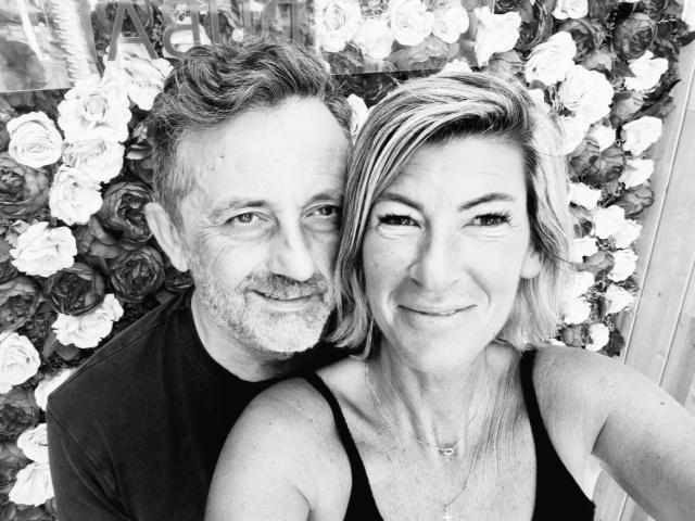 Black and white selfie of AngeelCouple, a middle-aged man with a gray beard and AngeelCouple, a woman with blonde hair and a black top, in front of roses.