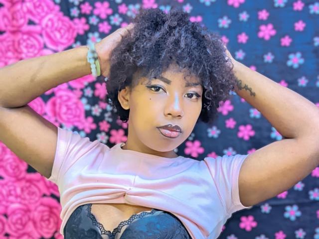 CamillaSexy with curly black hair, hands in her hair, wearing a pink shirt lifted to expose black lace bra, wearing green bracelet, looking at camera with pouty lips.