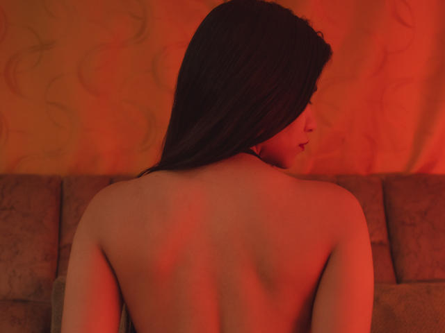 AnastasiiaS is topless with her back to the camera, showing her bare ass and smooth skin, with long dark hair and a red-orange light.