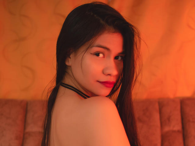 AnastasiiaS with long black hair, looking over her shoulder, showing her bare shoulder and smiling with red lipstick, warm orange light, and a soft brown cushioned seat.