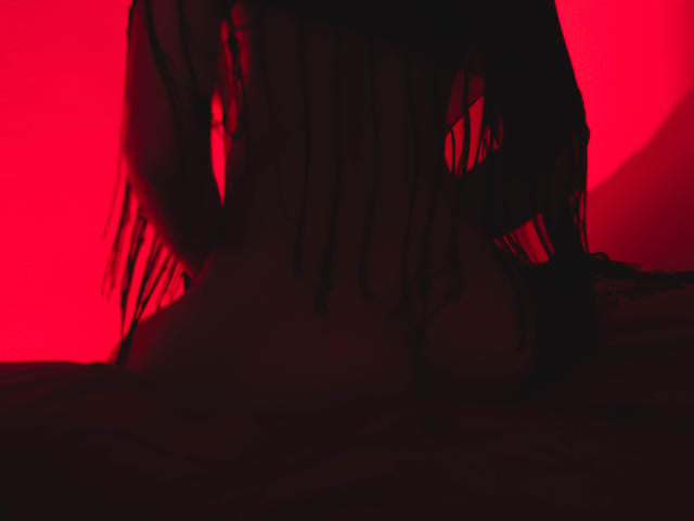 AnastasiiaS's dark, fringed scarf partially covers her naked, red-lit body, revealing her small, round tits and a hint of her ass, with her hands resting on a surface.